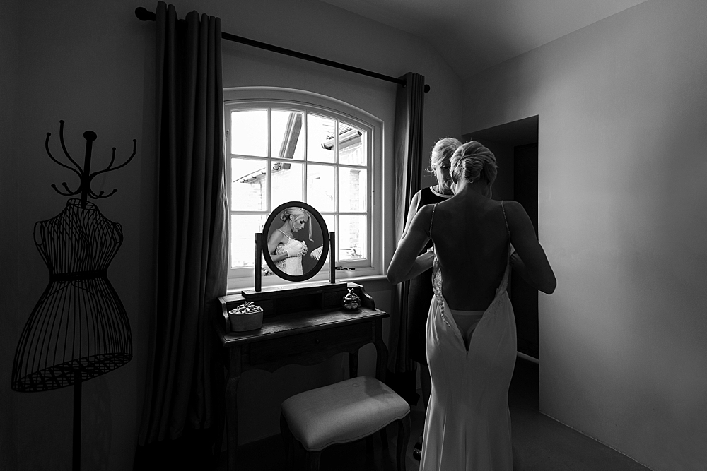 Black and white wedding photo of a bride-to-be at Southend Barns wedding venue in Chichester getting ready during bridal prep in the milking parlour and bridesmaid doing up her Enzoani wedding dress
